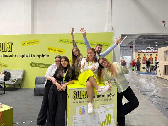 The SUPA team posing and smiling in front of their bright green booth at an event.